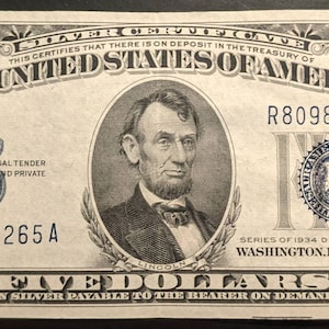 May include: A vintage five-dollar silver certificate from 1934. The bill features a portrait of Abraham Lincoln and the text "The United States of America." The bill is green and black with the number "5" in each corner.