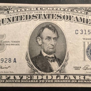 May include: A vintage five dollar bill featuring a portrait of Abraham Lincoln. The bill is printed in black and white with blue ink accents. The bill has the text "The United States of America" and "Five Dollars in Silver Payable to the Bearer on Demand".