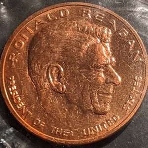 May include: A copper-coloured coin featuring a profile of Ronald Reagan, the 40th President of the United States, on the obverse. The coin reads "Ronald Reagan" and "President of the United States".