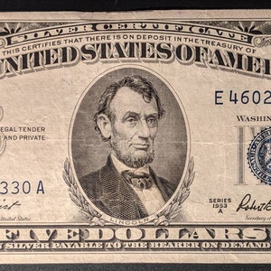 May include: A vintage five dollar bill featuring a portrait of Abraham Lincoln. The bill is dated 1953 and has the serial number E 46020330 A. The text "THE UNITED STATES OF AMERICA" is printed in bold letters at the top of the bill.