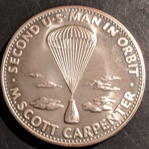 May include: Silver coin featuring the inscription "Second U.S. Man in Orbit M. Scott Carpenter" and an image of a parachute.