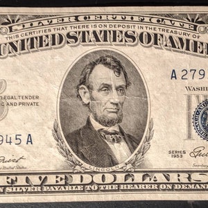 May include: A five dollar silver certificate from 1953 featuring a portrait of Abraham Lincoln. The serial number is A 27918945 A. The text "This certifies that there is on deposit in the Treasury of the United States of America" is printed at the top of the bill.