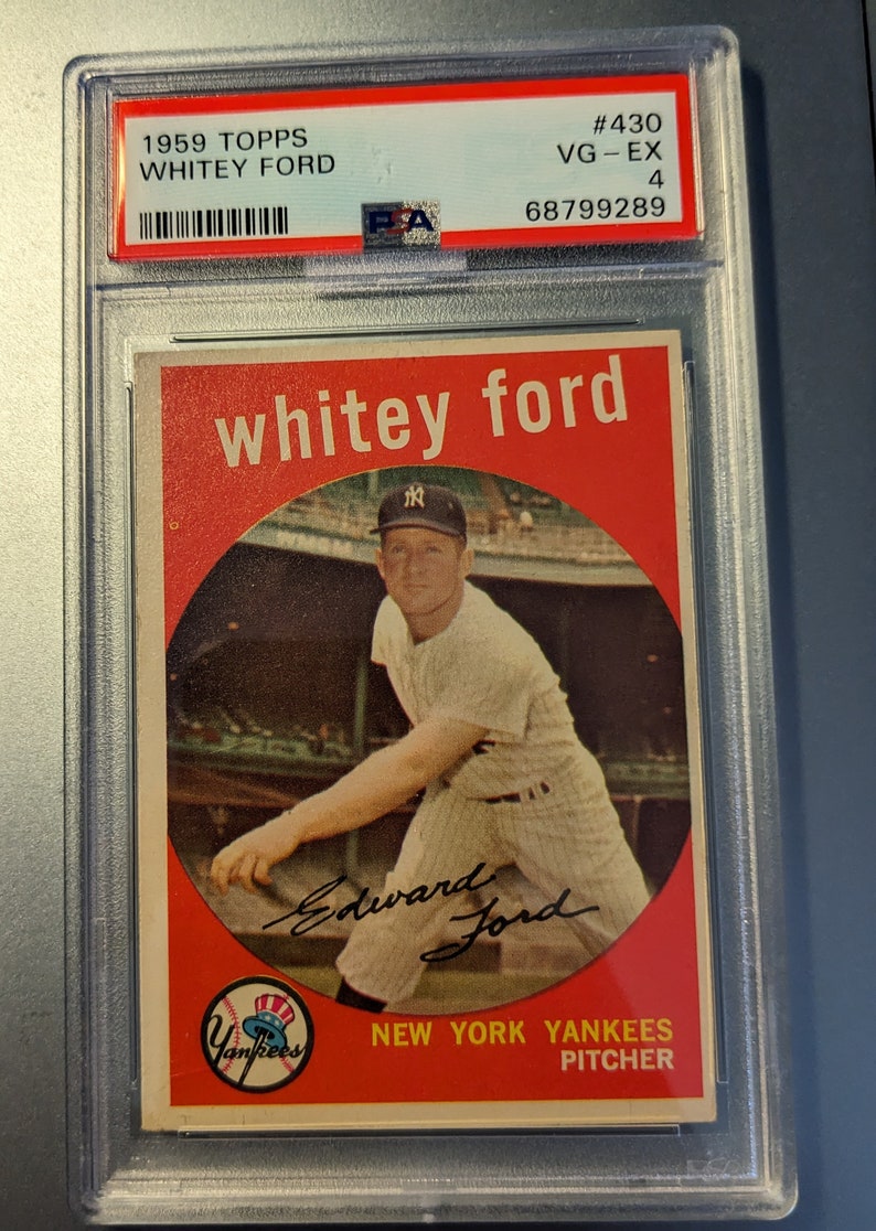 May include: A 1959 Topps baseball card featuring a photo of Whitey Ford, a pitcher for the New York Yankees. The card is graded VG-EX 4 and has a PSA authentication sticker. The card has a red background with a white circle in the center. The circle contains a photo of Whitey Ford pitching. The card also includes Whitey Ford's signature.