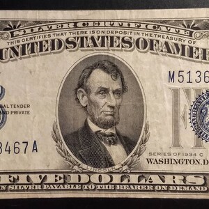 May include: A vintage five dollar silver certificate from 1934. The certificate features a portrait of Abraham Lincoln and the text "THE UNITED STATES OF AMERICA". The serial number is M51363467A.