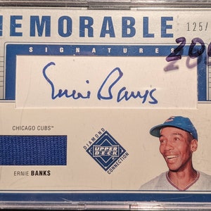 Può includere: Una carta di baseball con Ernie Banks dei Chicago Cubs. La carta è della serie Upper Deck Diamond Connection ed è numerata 125/150. La carta ha un pezzo di maglia blu e una firma di Ernie Banks.