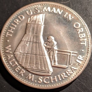 May include: A silver commemorative coin depicting Walter M. Schirra Jr., the third U.S. man in orbit, standing on a platform next to a space capsule. The coin reads "THIRD U.S. MAN IN ORBIT WALTER M. SCHIRRA JR."