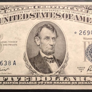 May include: A vintage five-dollar bill from the United States, featuring a portrait of Abraham Lincoln. The bill has intricate border designs and text that reads "The United States of America" and "Five Dollars". The bill is in shades of green and black.