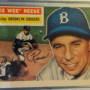May include: A vintage baseball card featuring Pee Wee Reese, a shortstop for the Brooklyn Dodgers. The card has a red and white border with the text "Pee Wee" Reese, shortstop Brooklyn Dodgers. The card features a color photo of Reese in a blue baseball cap with a white "B" on it. Reese is smiling and looking to the right. The card also features a black and white photo of Reese in a baseball uniform, crouching on the field. The card is signed in black ink by Reese.