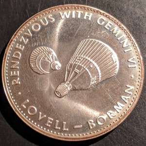 May include: A silver coin with the text "Rendezvous with Gemini" and "Lovell - Borman" around the edge. The coin features a depiction of two space capsules.