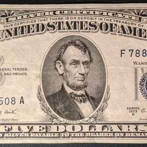 May include: A five dollar silver certificate from 1953 featuring a portrait of Abraham Lincoln. The certificate has the text "The United States of America" and "Five Dollars in Silver Payable to the Bearer on Demand".