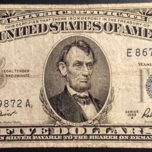 May include: A vintage five dollar bill featuring a portrait of Abraham Lincoln. The bill is dated 1953 and has the serial number E86739872 A. The text "THE UNITED STATES OF AMERICA" is printed in bold letters at the top of the bill.