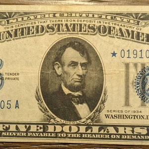 May include: A vintage five dollar bill from the United States of America, featuring a portrait of Abraham Lincoln and the text "THIS CERTIFIES THAT THERE IS ON DEPOSIT IN THE TREASURY OF THE UNITED STATES OF AMERICA FIVE DOLLARS IN SILVER PAYABLE TO THE BEARER ON DEMAND". The bill is dated "SERIES OF 1934 WASHINGTON, D.C.".
