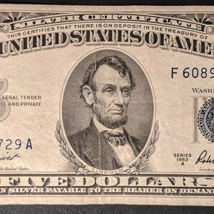 May include: A vintage five dollar bill featuring a portrait of Abraham Lincoln. The bill is in black and white with a blue seal and text that reads "The United States of America" and "Five Dollars in silver payable to the bearer on demand".