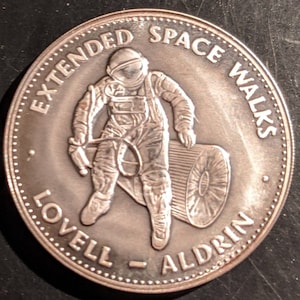 May include: A silver coin with a depiction of an astronaut in a space suit, holding a rolled-up object. The coin has the text "EXTENDED SPACE WALKS LOVELL - ALDRIN" around the edge.