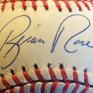 May include: A white baseball with red stitching and a blue signature that reads "Brian Rae".