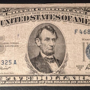 May include: A vintage five dollar bill featuring a portrait of Abraham Lincoln. The bill is in black and white with blue and red accents. The serial number is F 46864325 A. The bill is dated 1953.