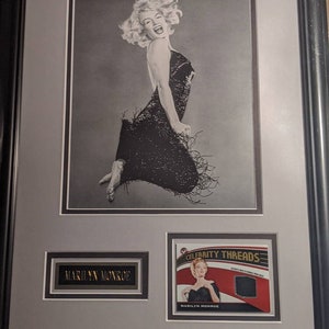 Puede incluir: Una fotografía en blanco y negro enmarcada de Marilyn Monroe con un vestido negro de lentejuelas. La foto está montada con una pequeña tarjeta con el texto "Celebrity Threads" y una muestra de tela. La tarjeta también incluye una fotografía en blanco y negro más pequeña de Marilyn Monroe.
