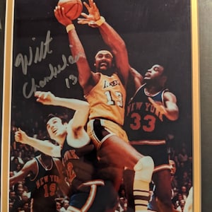 Puede incluir: Fotografía vintage enmarcada de un partido de baloncesto con Wilt Chamberlain con el uniforme de los Lakers. La imagen muestra a los jugadores en acción, con Chamberlain alcanzando el balón. La foto está firmada y numerada.