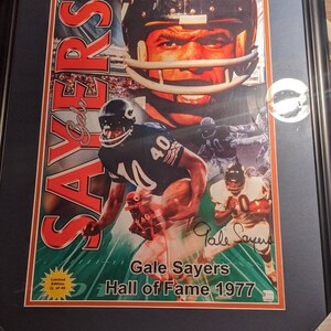 Puede incluir: Un póster enmarcado de Gale Sayers, un ex jugador de fútbol americano. El póster presenta un collage de imágenes de Sayers con su uniforme de los Chicago Bears, incluyendo un primer plano de su rostro. El póster se titula "Gale Sayers Hall of Fame 1977" y está numerado "2 de 40".