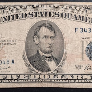 May include: A vintage five dollar silver certificate featuring a portrait of Abraham Lincoln. The certificate is dated 1953 and has the serial number F 34321048 A. The text "THE UNITED STATES OF AMERICA" is printed in bold at the top of the certificate.
