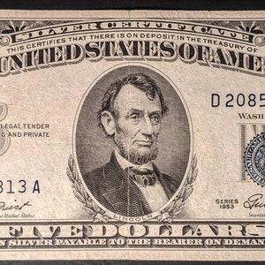 May include: A vintage five dollar silver certificate featuring a portrait of Abraham Lincoln. The certificate is dated 1953 and has the serial number D20857813A. The text "THE UNITED STATES OF AMERICA" is printed in bold at the top of the certificate.