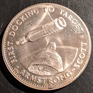 May include: A silver coin with the text "First  Docking to Target Armstrong-Scott" around the edge. The coin features a depiction of a spacecraft docking with a larger spacecraft.