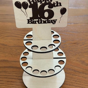 Peut inclure: Pr&eacute;sentoir en bois pour un 16e anniversaire. Le haut porte l'inscription "Happy 16th Birthday" avec des ballons. Le pr&eacute;sentoir comporte trois niveaux avec des d&eacute;coupes circulaires pour contenir des objets. Le bois est de couleur naturelle.