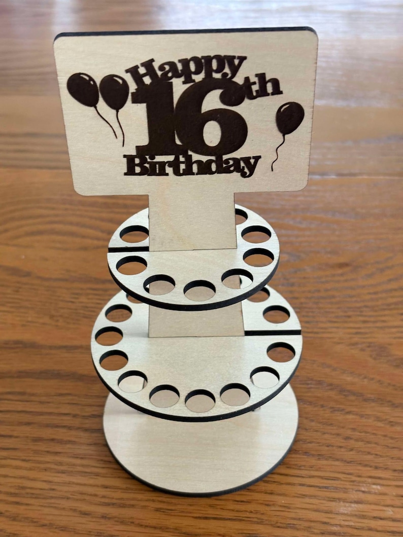 Peut inclure: Pr&eacute;sentoir en bois pour un 16e anniversaire. Le haut porte l'inscription "Happy 16th Birthday" avec des ballons. Le pr&eacute;sentoir comporte trois niveaux avec des d&eacute;coupes circulaires pour contenir des objets. Le bois est de couleur naturelle.