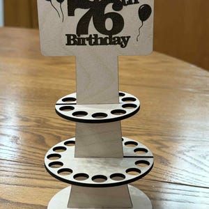 Peut inclure: Pr&eacute;sentoir d'anniversaire en bois avec l'inscription "Happy 76th Birthday" et des ballons. Le pr&eacute;sentoir comporte trois niveaux avec des d&eacute;coupes circulaires, probablement pour contenir des objets. Le design est en bois clair avec des accents noirs.