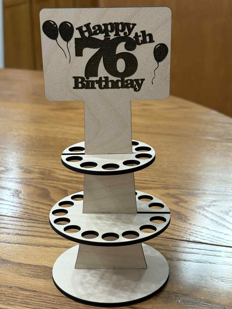 Peut inclure: Pr&eacute;sentoir d'anniversaire en bois avec l'inscription "Happy 76th Birthday" et des ballons. Le pr&eacute;sentoir comporte trois niveaux avec des d&eacute;coupes circulaires, probablement pour contenir des objets. Le design est en bois clair avec des accents noirs.