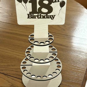 Peut inclure: Pr&eacute;sentoir en bois pour un 18e anniversaire. Le panneau indique "Happy 18th Birthday" avec des ballons. Le pr&eacute;sentoir comporte trois niveaux circulaires avec plusieurs trous, con&ccedil;us pour contenir des articles. Fabriqu&eacute; en bois clair.