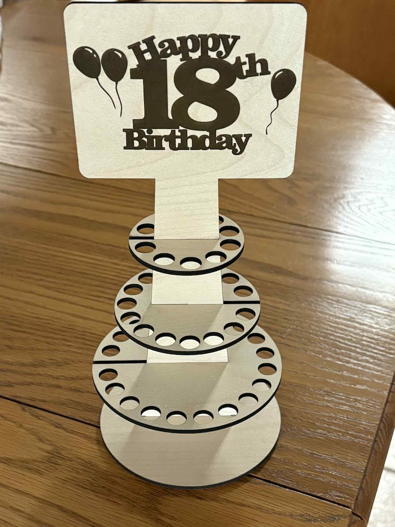 Peut inclure: Pr&eacute;sentoir en bois pour un 18e anniversaire. Le panneau indique "Happy 18th Birthday" avec des ballons. Le pr&eacute;sentoir comporte trois niveaux circulaires avec plusieurs trous, con&ccedil;us pour contenir des articles. Fabriqu&eacute; en bois clair.