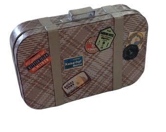 May include: A rectangular suitcase with a tan and brown woven pattern, featuring a silver handle and two vertical light-colored straps. The surface is decorated with various travel-themed stickers, including labels for Hotel Hessler, Kaiserhof Berlin, and The Kamenoi Hotel.