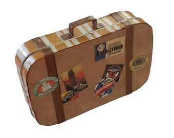 May include: A rectangular suitcase with a tan body and brown vertical straps. The top features white and tan stripes. It is decorated with various vintage-style travel stickers, including labels for Hotel D'Angleterre Geneve and Hotel Mondial.
