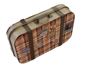 May include: A rectangular metal suitcase with a tan and brown plaid pattern, featuring a top handle, two dark brown vertical straps, and various vintage-style travel stickers depicting scenes like a camel and hotel logos on the front surface.