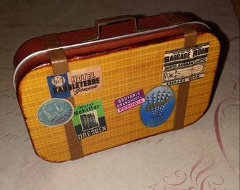 May include: A miniature brown suitcase with a metal handle and straps. The suitcase is decorated with vintage travel stickers, including "Hotel Angleterre Genive", "Hotel Schiller Dresden", "Brufanis Grandhotel Perugia", and "Baggage Room North German Lloyd".