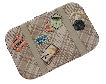 May include: A rectangular metal tin featuring a beige and brown plaid pattern with two light brown straps. The surface is decorated with various vintage-style travel stickers, including labels for Hotel Hessler, Kaiserhof Berlin, Kamenoi Hotel, and Royal York.