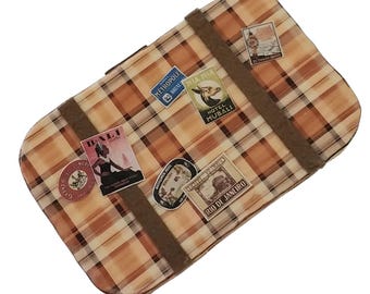 May include: A rectangular suitcase featuring a tan and brown plaid pattern with two dark brown horizontal straps. The surface is decorated with several vintage-style travel stickers, including labels for Bali, Hotel Mubali, and Rio de Janeiro.