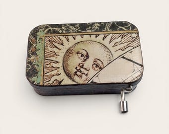 May include: A rectangular music box with a vintage sun illustration on the lid. The box has a black base and a silver winding key. The lid features a detailed sun face with rays and map-like elements.
