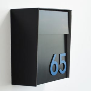May include: A black mailbox with a hinged top and a blue number 65. The mailbox is mounted on a white wall. The design is modern and minimalist.