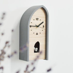 May include: A gray and wood-tone wall clock with a bird-shaped cuckoo. The clock has a unique arched design with black hands and hour markers. The clock is mounted on a white wall.