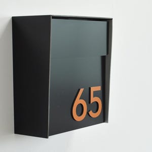 May include: A modern black mailbox with a rectangular design. The mailbox features the number "65" in a contrasting orange color. The mailbox is mounted on a white wall.