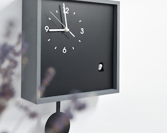 Könnte beinhalten: Eine quadratische Wanduhr mit dunkelgrauem Rahmen und schwarzem Zifferblatt. Die Uhr verfügt über weiße Stundenmarkierungen, weiße Zeiger und eine kleine weiße Vogelfigur, die durch einen Ausschnitt unten rechts sichtbar ist.