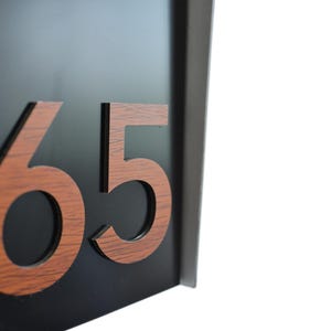 Könnte beinhalten: Schwarzes Hausnummernschild mit der Nummer 65 in Holzmaserung. Die Zahlen sind vor einem schwarzen Hintergrund erhaben. Das Schild ist rechteckig und hat ein modernes Design.