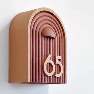 May include: A wall-mounted address plaque in a warm, earthy tone, featuring the number "65". The plaque has a rounded top and a carved, arched design with vertical lines and a half-circle detail. The design is reminiscent of a rainbow.