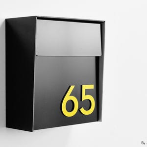 May include: A black mailbox with a yellow number 65. The mailbox is rectangular with a top flap. The numbers are large and easy to read. The mailbox is mounted on a white wall.