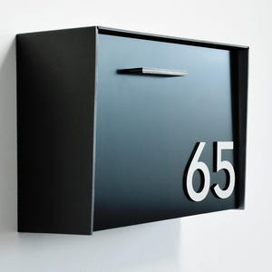 Può includere: Una moderna cassetta delle lettere nera con il numero 65 in argento. La cassetta delle lettere ha un design elegante e rettangolare con una piccola maniglia argentata orizzontale. È montata su un muro bianco.