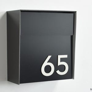 May include: A black mailbox with the number 65 in white. The mailbox is mounted on a white wall. The design is modern and minimalist.