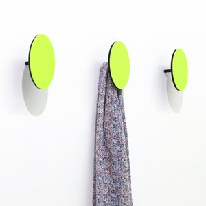 May include: Three round wall hooks with a black rim and a bright green center. The hooks are mounted on a white wall. A patterned scarf is hanging from one of the hooks.