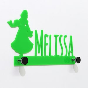Könnte beinhalten: Grüner Acryl-Wandhaken mit dem Namen "Melissa" und einer Silhouette einer Frau. Der Haken hat zwei silberne Haken und ist an einer weißen Wand montiert.